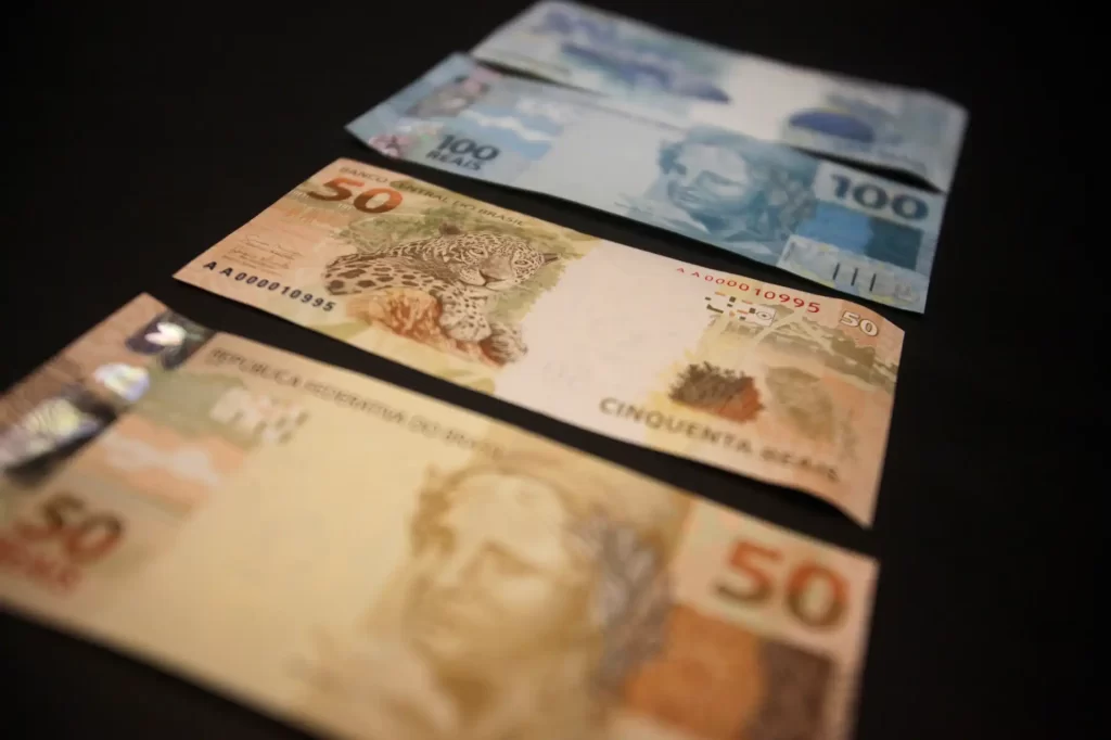 Imagem de notas de dinheiro de diferentes valores, incluindo uma nota de cinquenta e uma de cem reais, dispostas sobre uma superfície escura, destacando variedades de cédulas brasileiras.