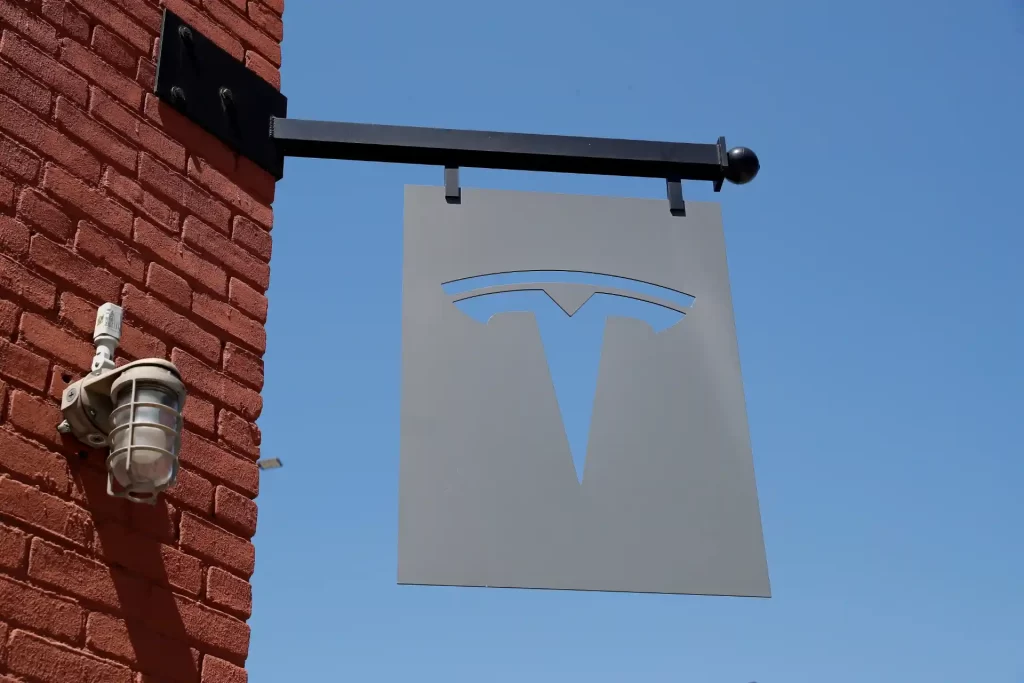 Placa de sinalização com o logotipo da Tesla em um fundo de céu azul. A placa está presa a uma parede de tijolos vermelhos.
