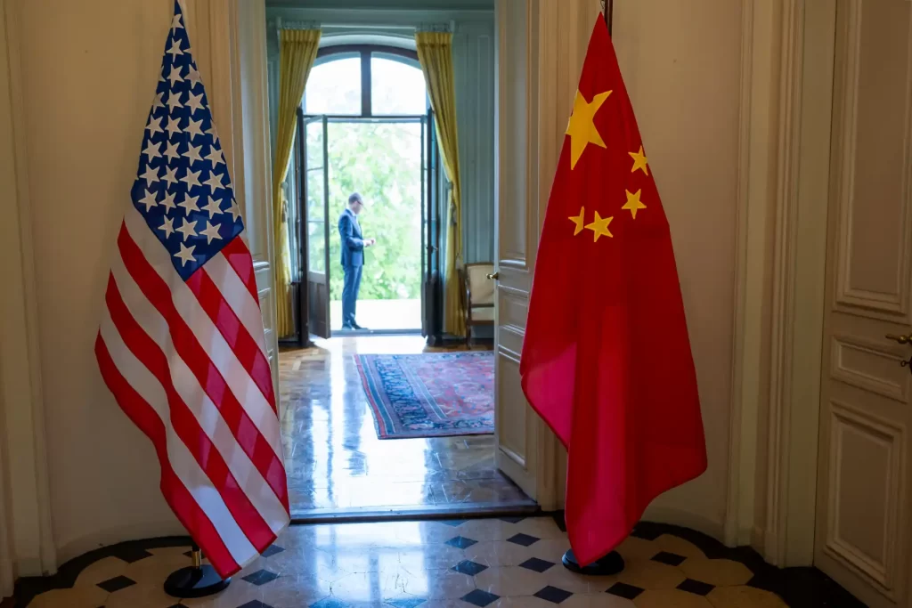 Reunião diplomática entre representantes dos Estados Unidos e China, com as bandeiras dos dois países em destaque e um homem ao fundo, apresentando ambiente formal e institucional.