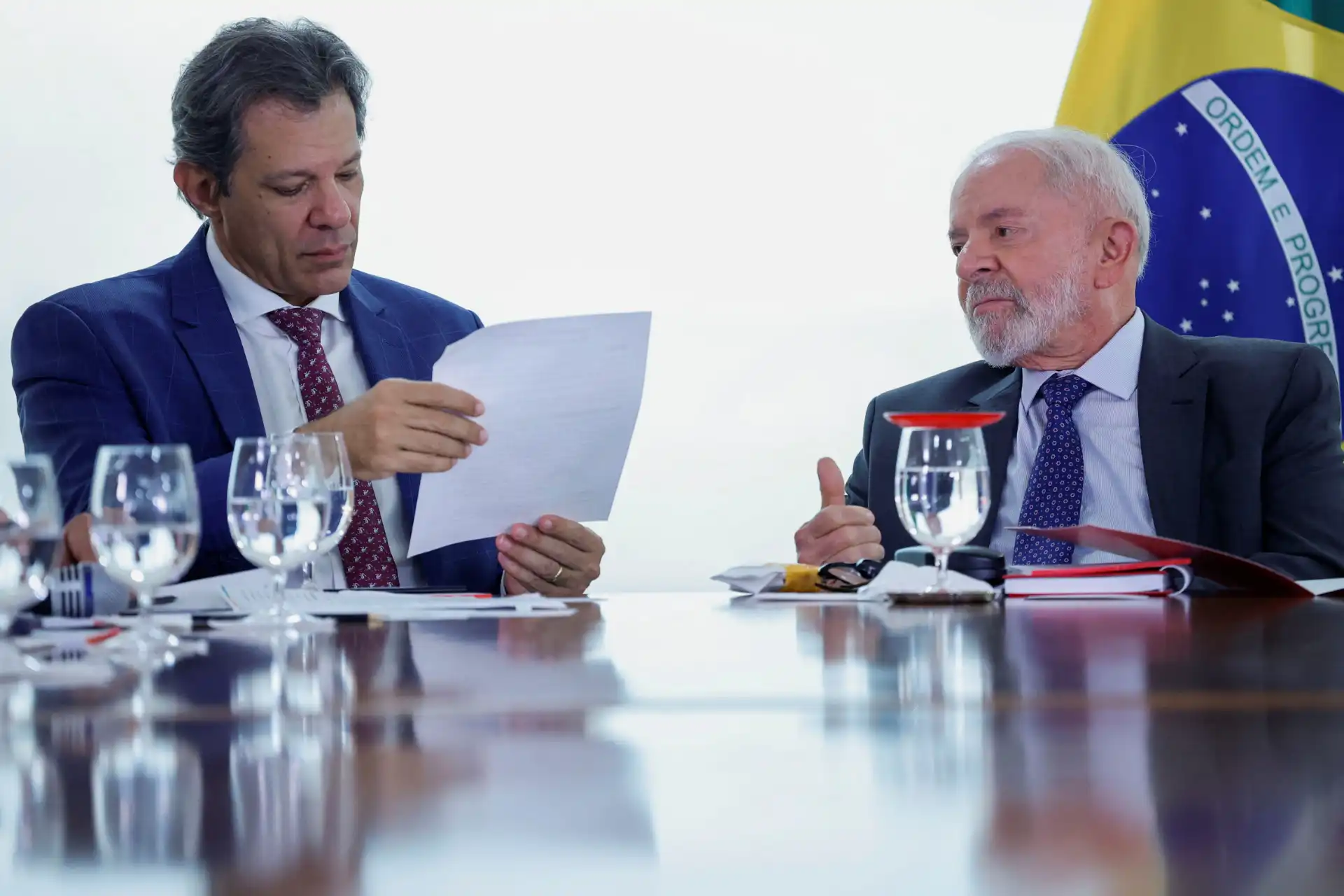 Reunião entre políticos brasileiros, com um deles segurando documentos, em ambiente formal com bandeira do Brasil ao fundo.