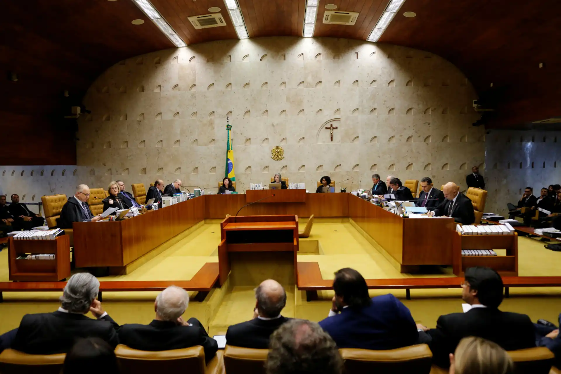 Sessão do Supremo Tribunal Federal no Brasil, com ministros e advogados presentes, em ambiente formal e com decoração moderna, destaque para a bandeira nacional e cruz na parede ao fundo.