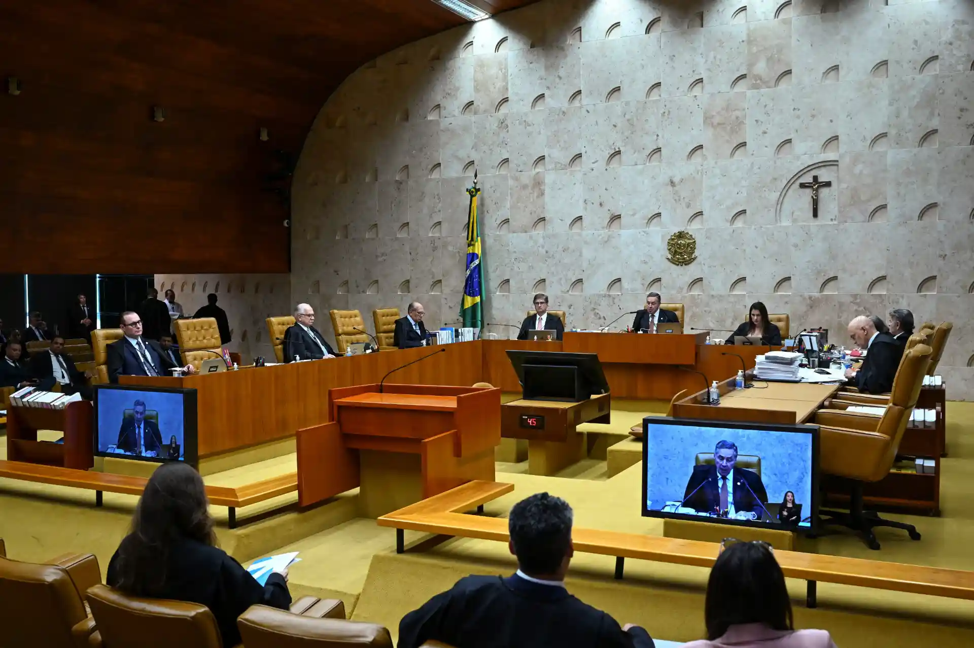 Sessão do tribunal ou assembleia legislativa com membros usando terno e computadores, uma sala elegante com parede de pedra, cruz e bandeira brasileira ao fundo.