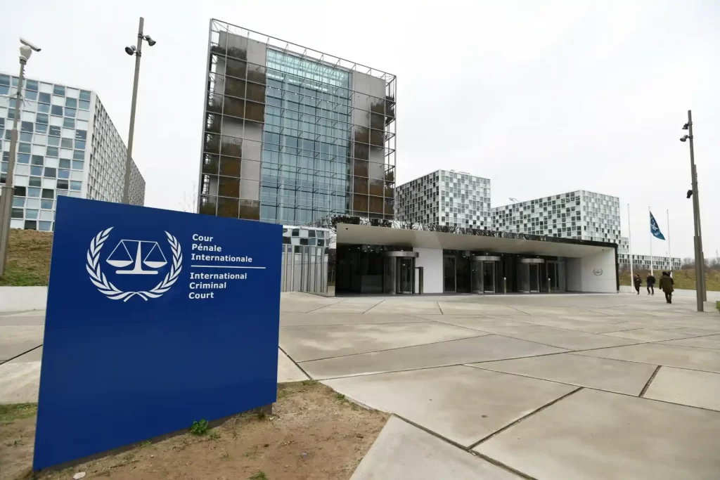 Imagem do Tribunal Penal Internacional localizado em Haia, Holanda, com sua fachada moderna de vidro e uma placa azul com símbolo de balança de justiça.