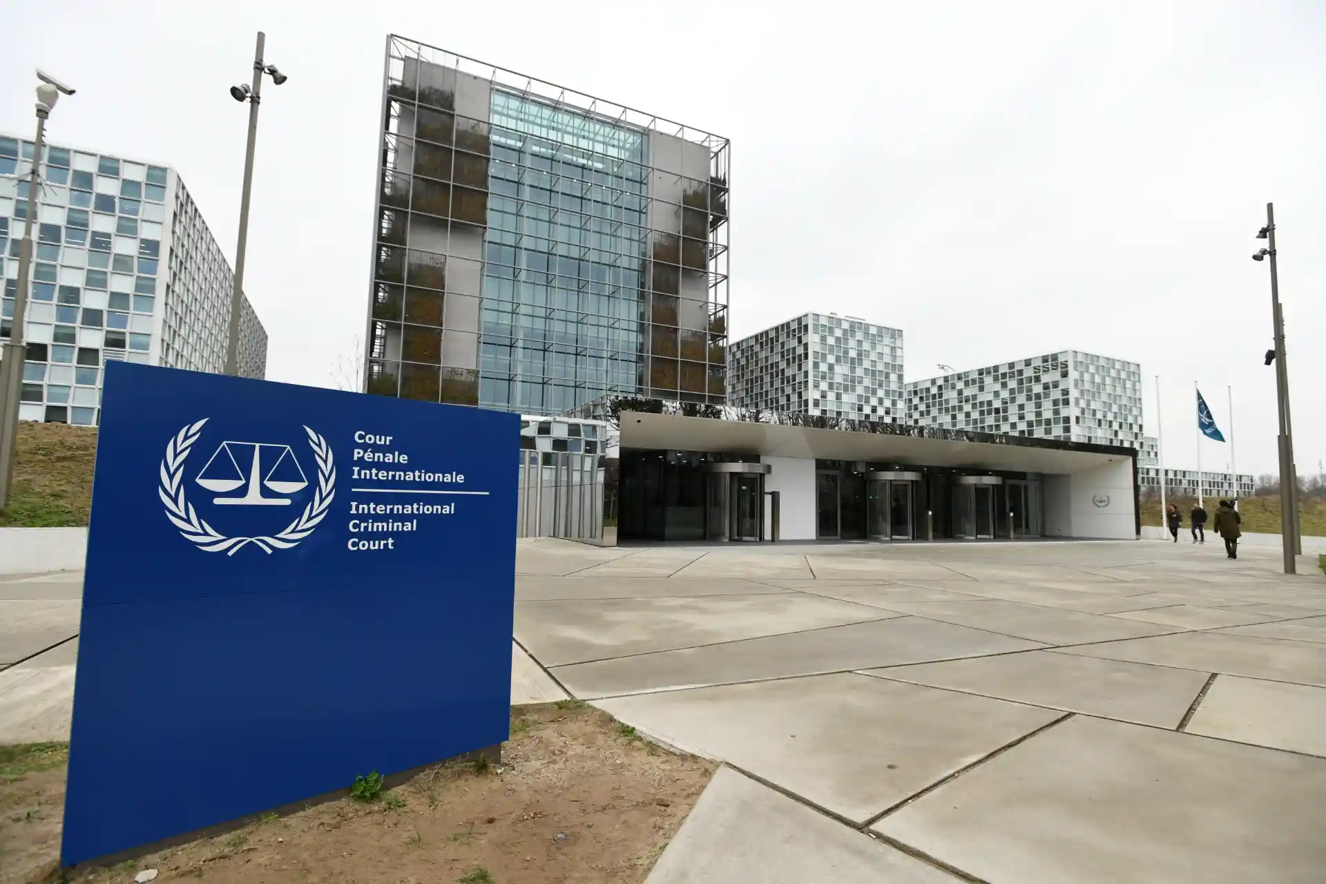 Imagem do Tribunal Penal Internacional localizado em Haia, Holanda, com sua fachada moderna de vidro e uma placa azul com símbolo de balança de justiça.