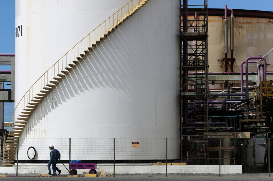 Imagem de uma refinaria de petróleo com torres de armazenamento e tubos, destacando a infraestrutura industrial e operários trabalhando no local.
