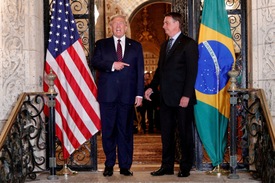Imagem de Donald Trump e um diplomata brasileiro conversando na entrada de um edifício com bandeiras dos Estados Unidos e do Brasil ao fundo. O cenário é elegante e oficial, refletindo uma reunião diplomática importante.