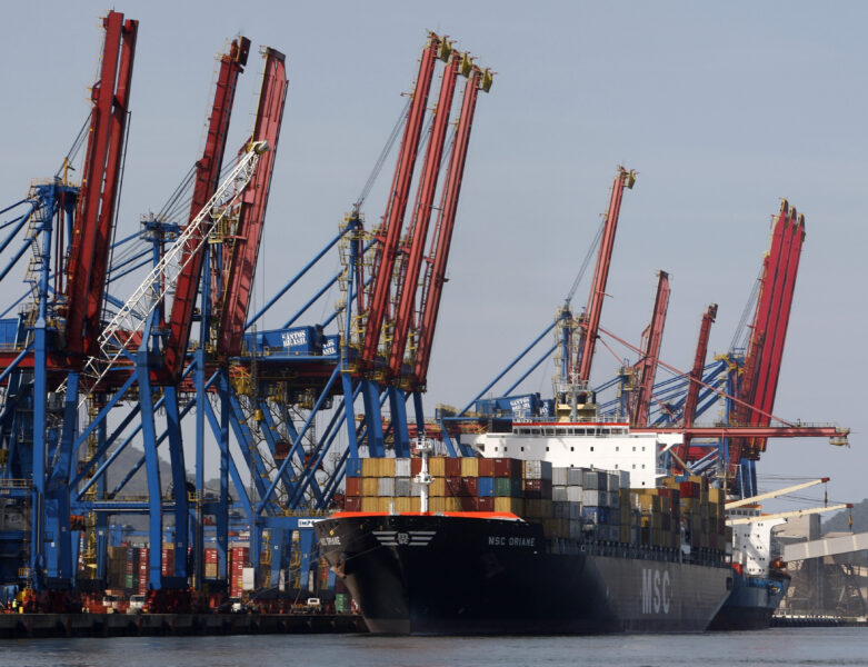 Imagem de um porto de carga com contêineres e guindastes gigantes em operação, destacando a atividade de transporte marítimo de cargas.