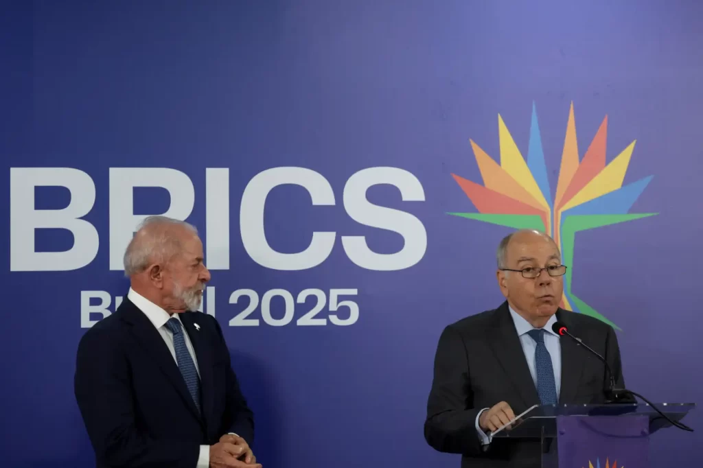 Duas pessoas em uma conferência na BRICS 2025, com um fundo com logo do BRICS e uma árvore estilizada multicolorida, um deles falando ao microfone.