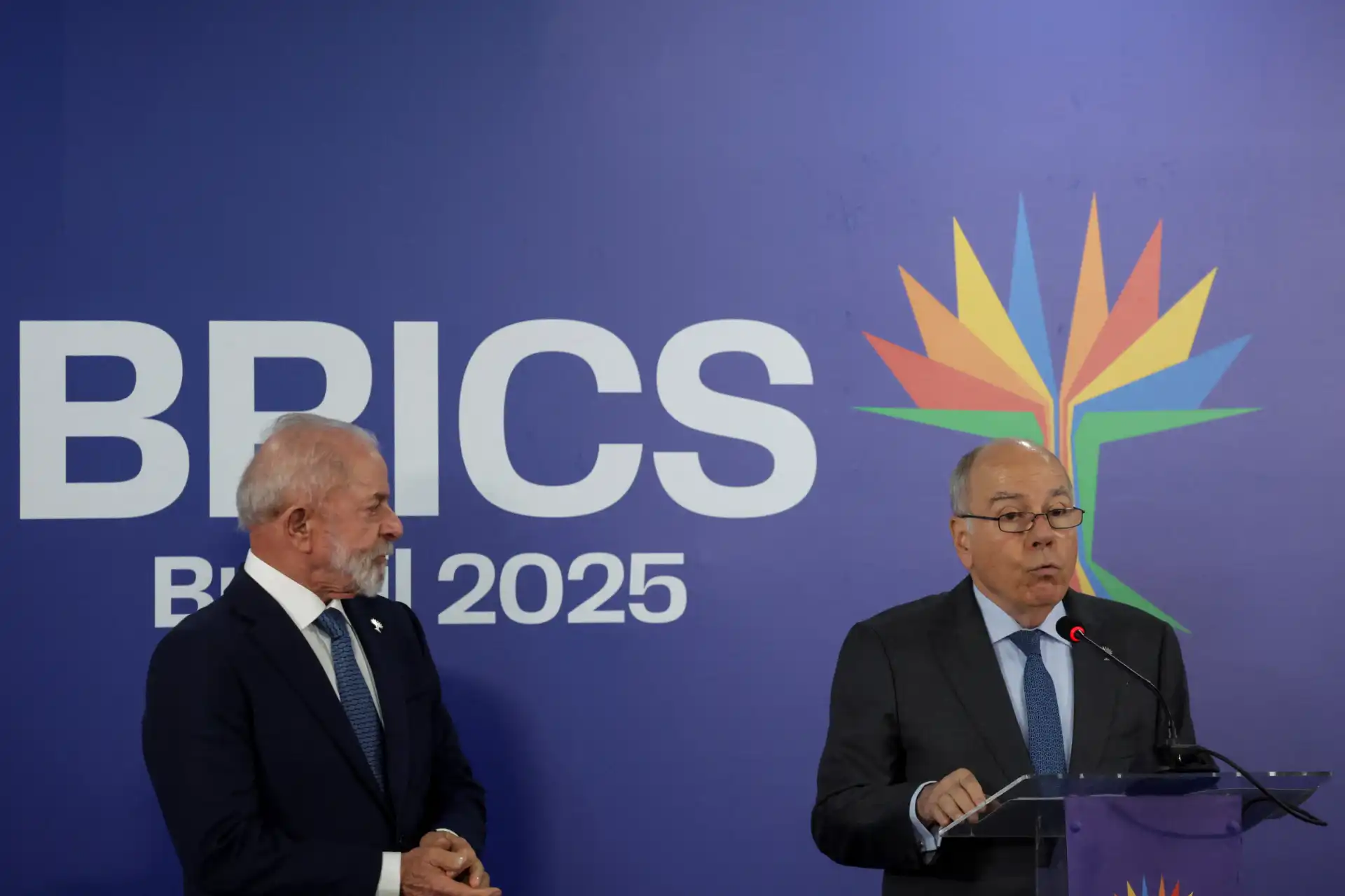 Duas pessoas em uma conferência na BRICS 2025, com um fundo com logo do BRICS e uma árvore estilizada multicolorida, um deles falando ao microfone.