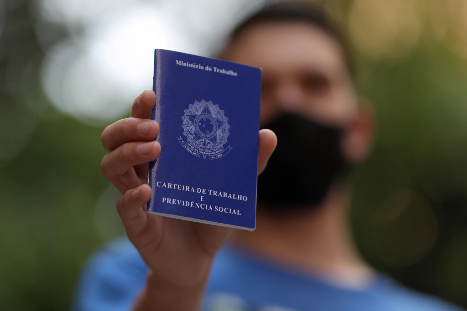 Pessoa segurando uma carteira de trabalho e previdência social do Ministério do Trabalho do Brasil, com máscara preta, ao ar livre.