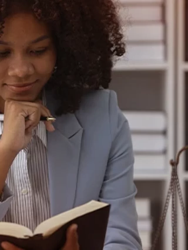 Mulher negra lendo um livro de forma pensativa em um escritório moderno, com estantes ao fundo e ambiente organizado, ideal para estudos ou leitura.