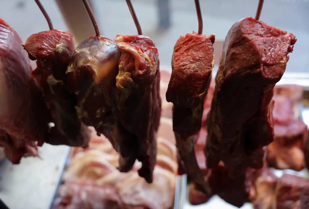 Cubos de carne bovina pendurados e prontos para preparo, ideal para churrasco ou assados, em ambiente de açougue bem iluminado.
