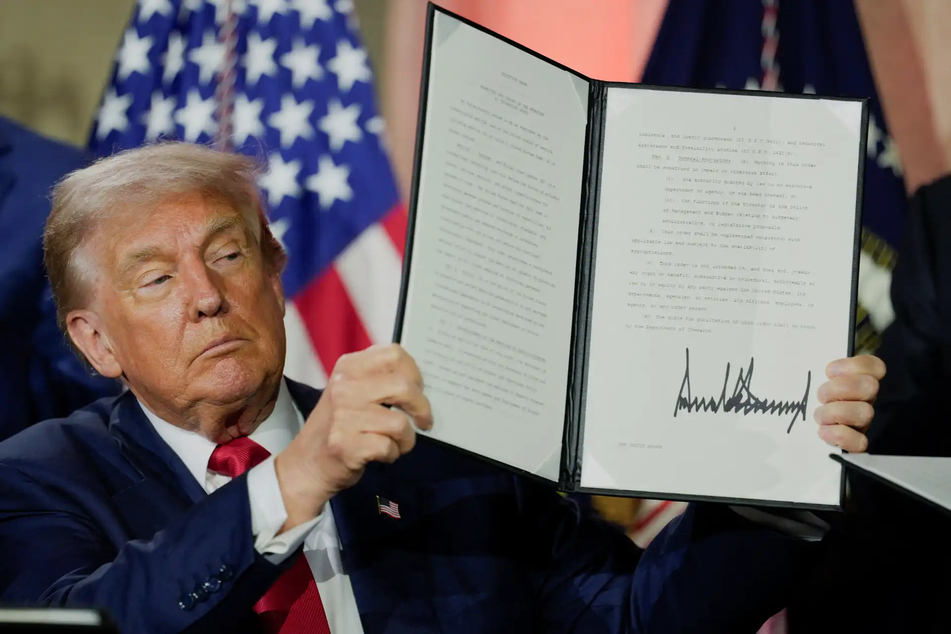 Imagem de Donald Trump segurando uma cópia de um documento assinado, durante evento oficial, com bandeiras ao fundo, transmitindo autoridade e assinatura de importante documento.