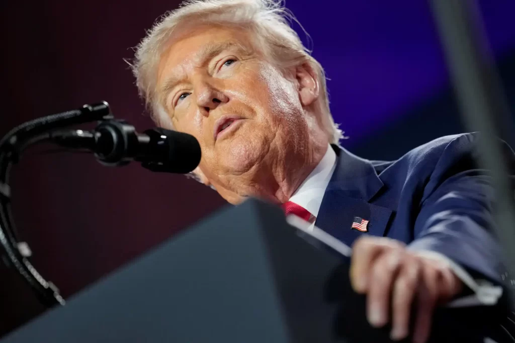 Imagem de Donald Trump falando em um microfone durante um discurso público, destacando sua expressão facial e traje formal.