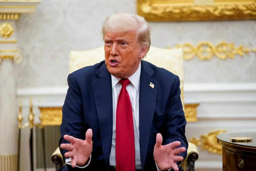 Imagem de Donald Trump sentado em uma sala decorada com detalhes dourados, usando terno azul, camisa branca e gravata vermelha, gesticulando enquanto fala.