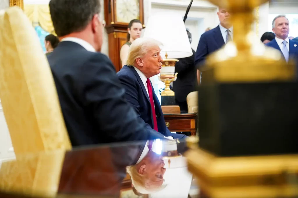 Trump anuncia armas para Ucrânia, alerta Rússia sobre "tarifas severas" se guerra continuar Imagem de Donald Trump sentado em reunião, rodeado por pessoas, em uma sala decorada de luxo, com foco na sua expressão e na atmosfera formal.