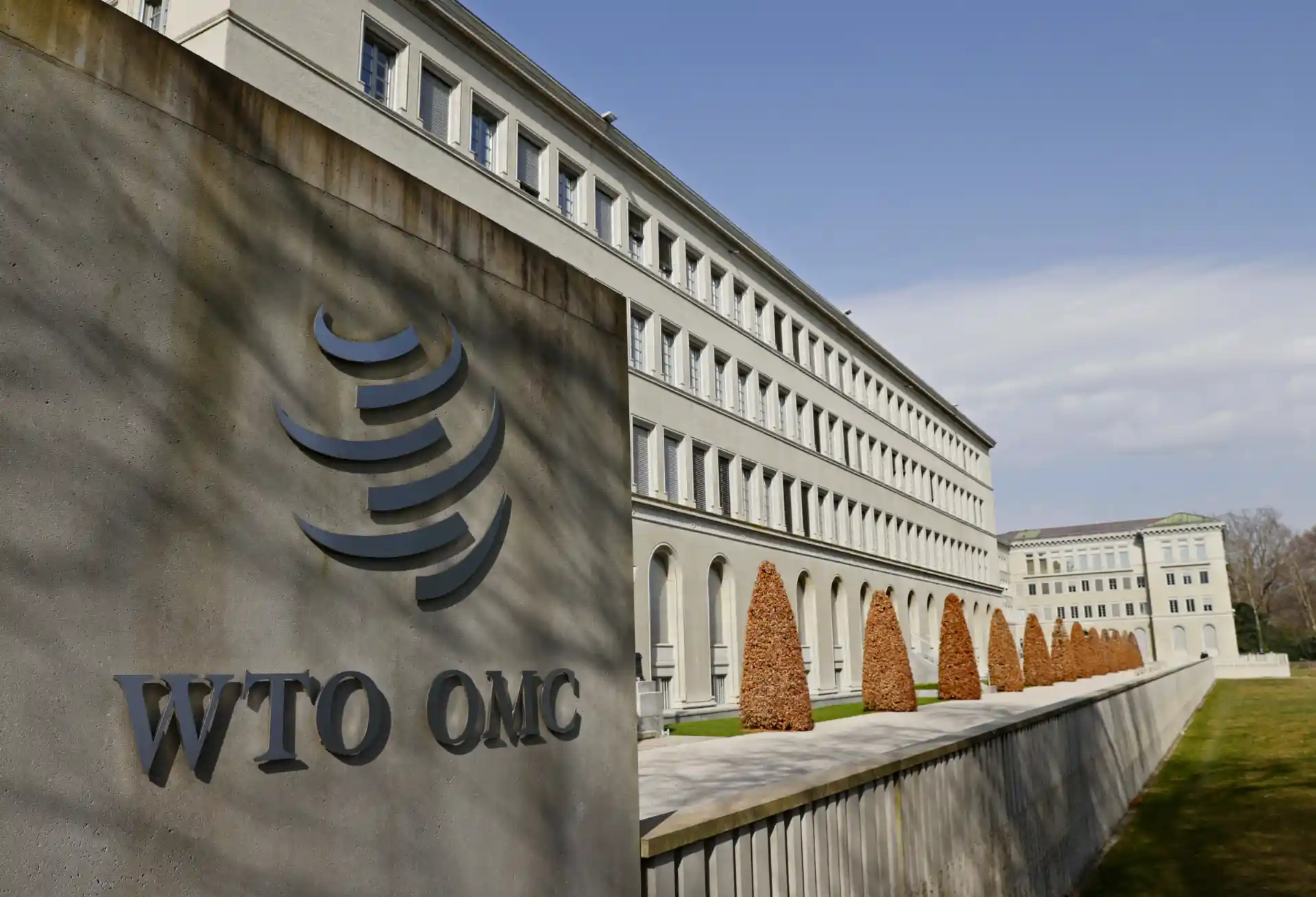Edifício da Organização Mundial do Comércio (OMC) em Genebra, Suíça, com destaque para o logo da OMC na parede de frente ao prédio, entre árvores e uma área verde bem cuidada.