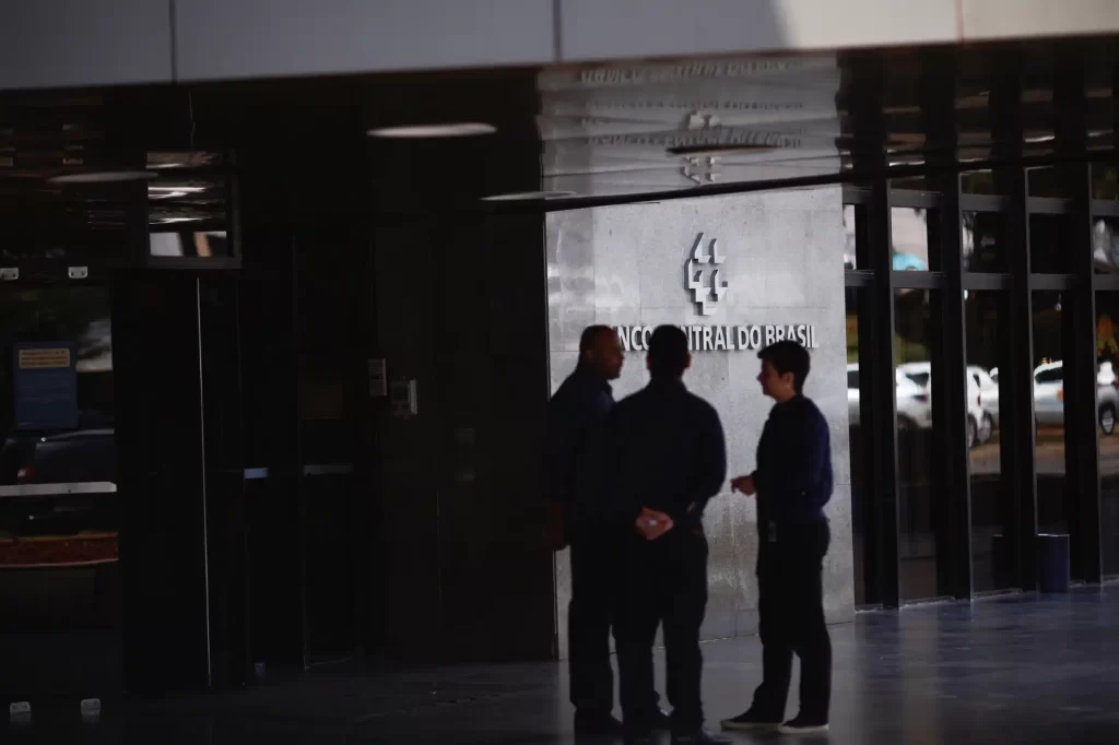 Imagem mostra três homens discutindo na entrada do Banco Central do Brasil, edificio moderno de fachada de vidro e símbolo do banco ao fundo, ambiente corporativo.
