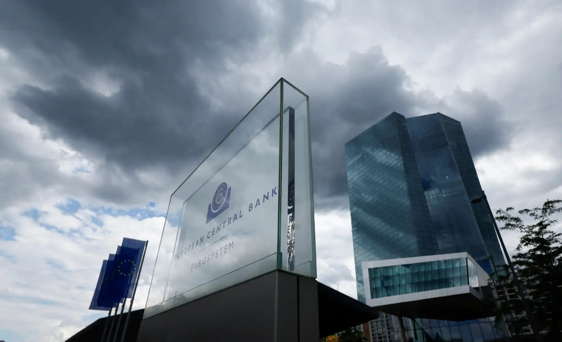 Imagem do European Central Bank com seu logotipo em destaque na fachada do edifício, sob céu nublado, em um dia cinza, em uma cidade europeia moderna.