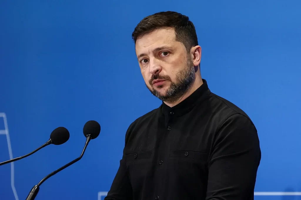 Homem em uma coletiva de imprensa, falando com seriedade, com fundo azul. Está vestido com camisa preta de manga longa.