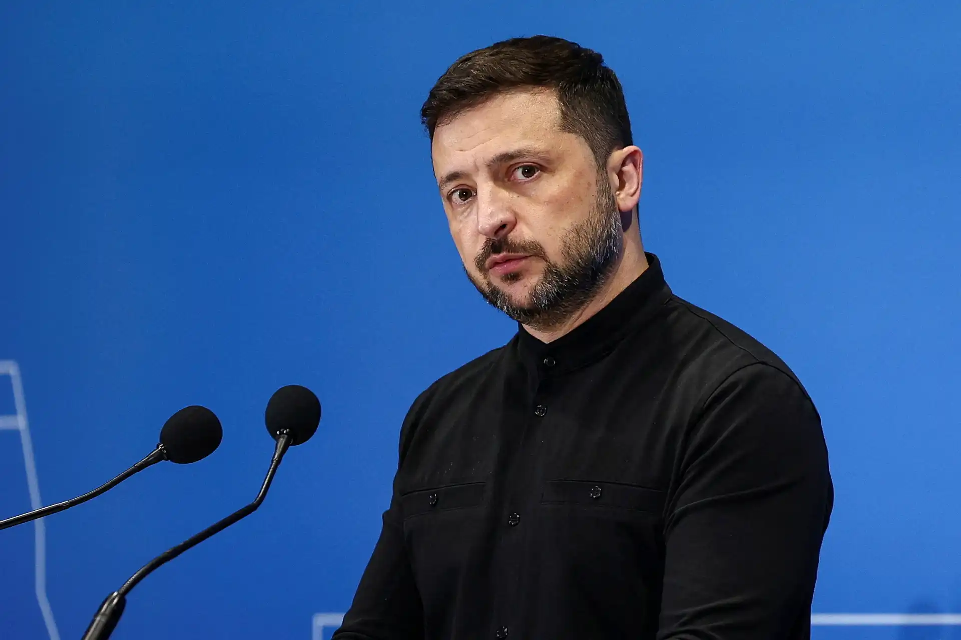 Homem em uma coletiva de imprensa, falando com seriedade, com fundo azul. Está vestido com camisa preta de manga longa.