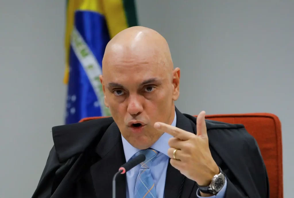 Imagem de um homem sério e confiante, vestido com terno e gravata, durante uma reunião ou discurso oficial, com bandeiras ao fundo.