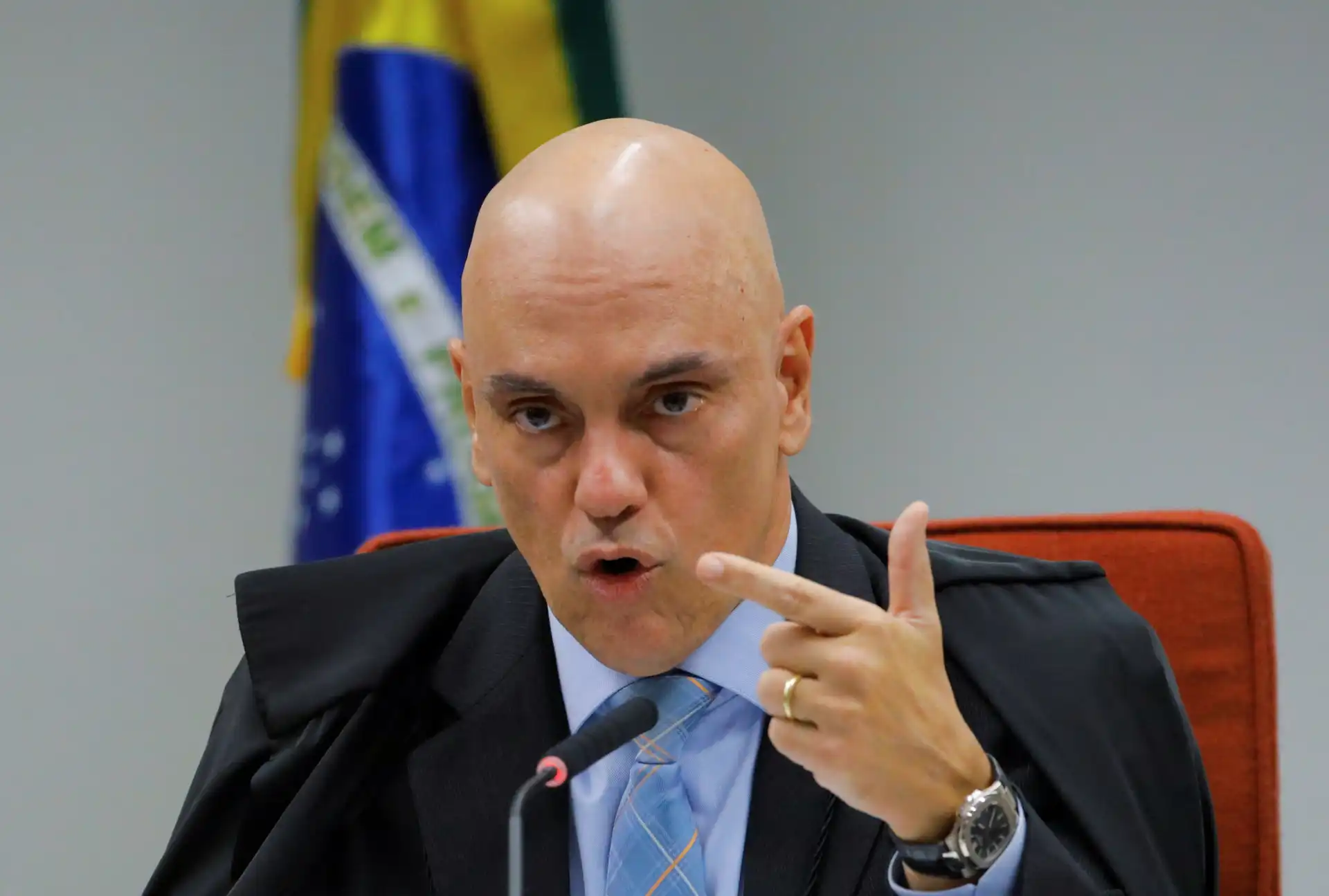 Imagem de um homem sério e confiante, vestido com terno e gravata, durante uma reunião ou discurso oficial, com bandeiras ao fundo.