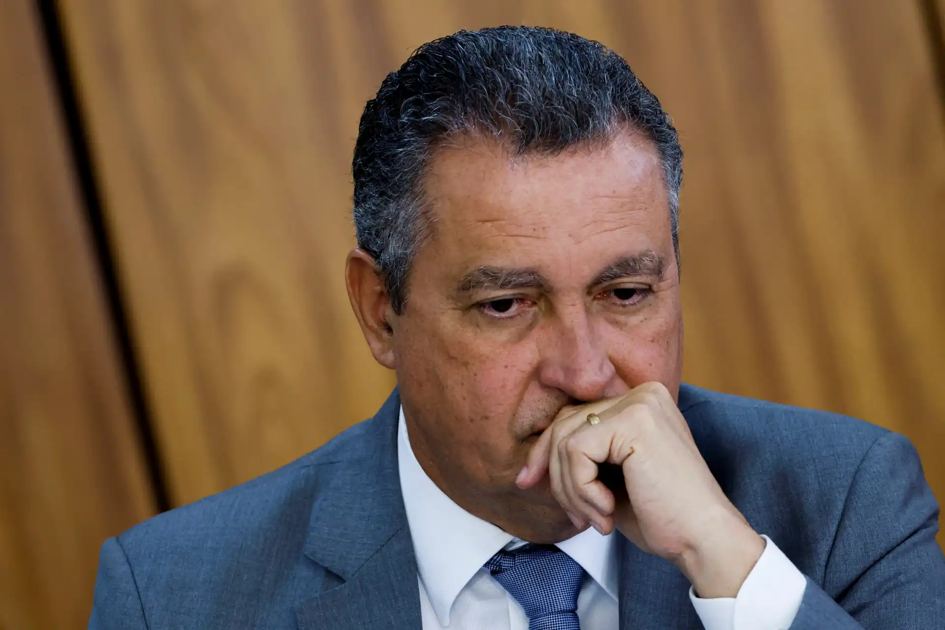 Imagem de um homem idoso com expressão pensativa, vestindo terno azul, sentado em um ambiente de madeira, representando reflexão ou preocupação.