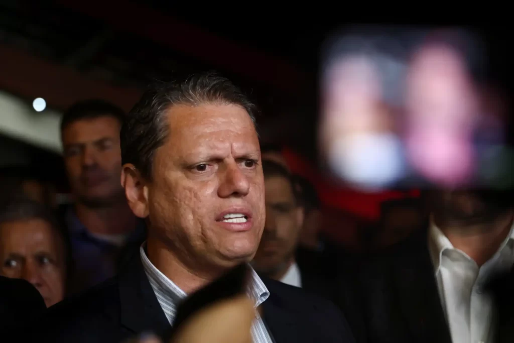 Homem expressando surpresa ou confusão, cercado por pessoas em um ambiente interno, possivelmente durante um evento ou conferência, foco principal no rosto.