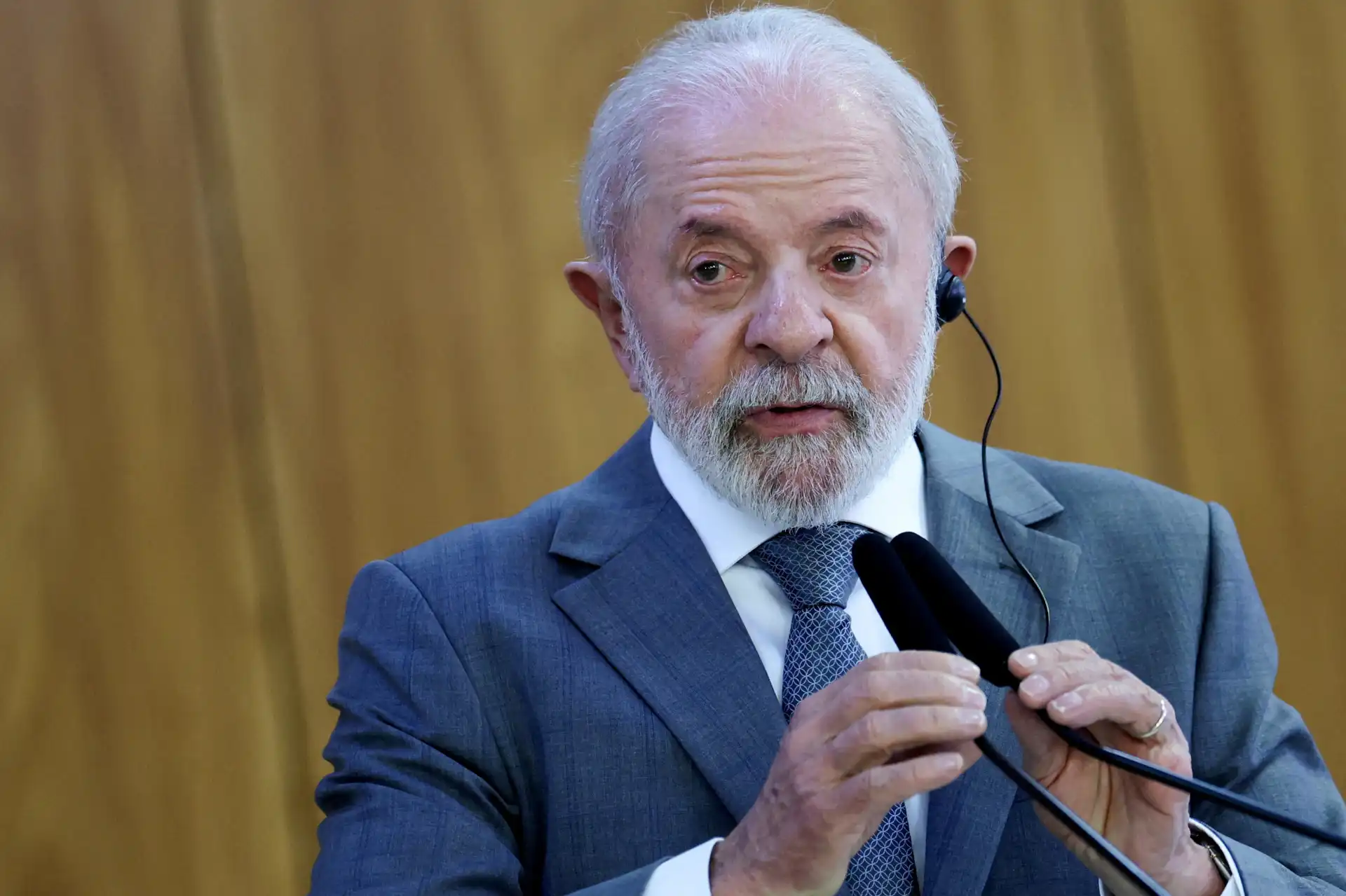 Homem de terno azul, cabelo branco e barba, falando em um microfone durante uma apresentação ou palestra com fundo de madeira.