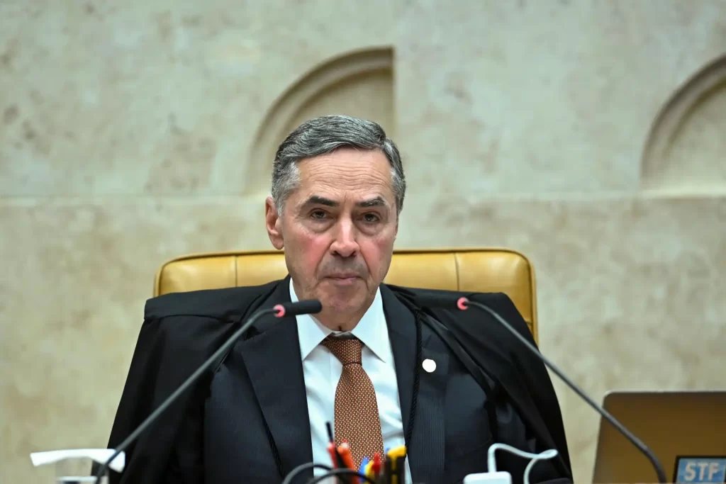 Imagem de um juiz sentado na mesa de tribunal, usando toga preta, com expressão séria. Fundo beige e painel na parede.