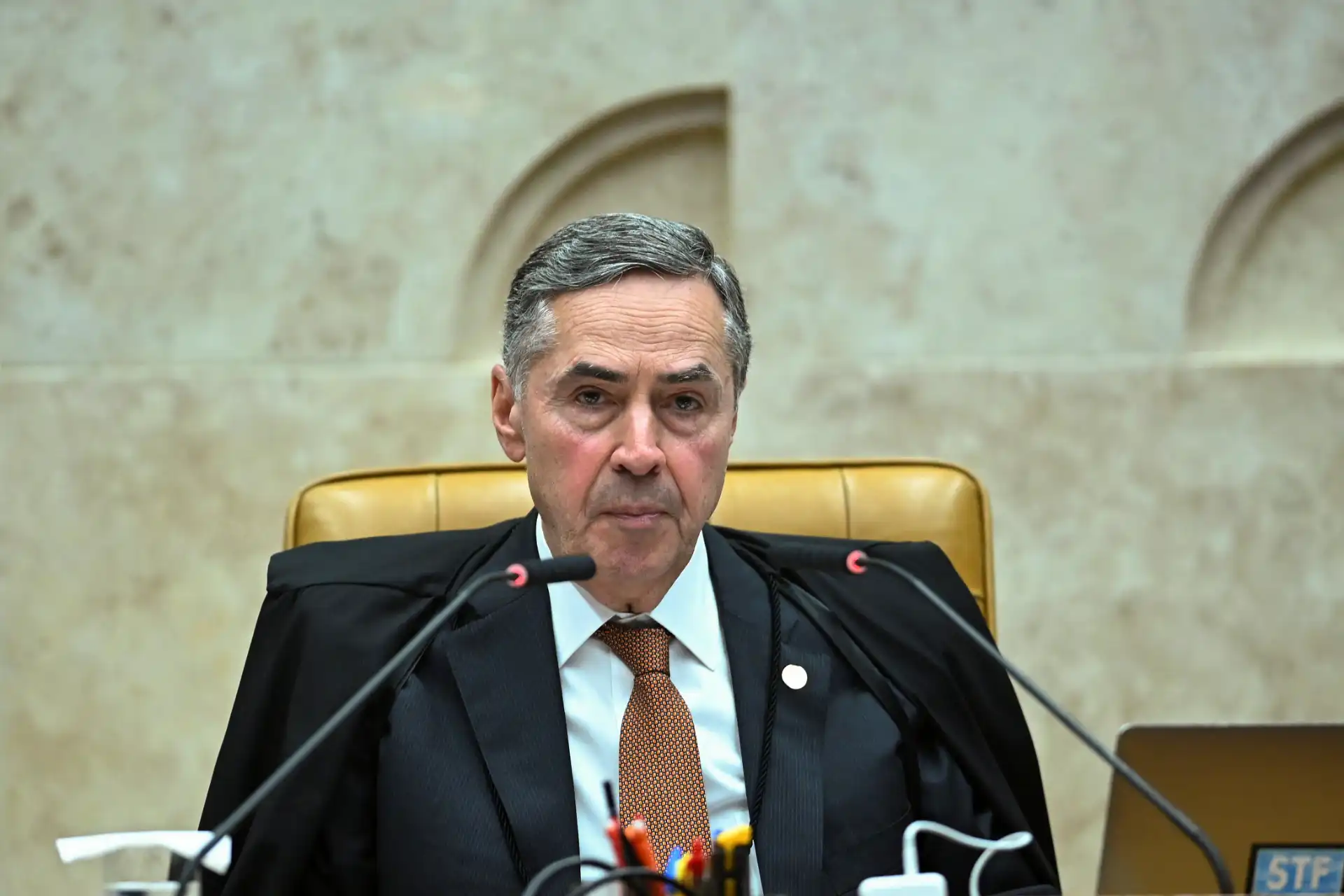 Imagem de um juiz sentado na mesa de tribunal, usando toga preta, com expressão séria. Fundo beige e painel na parede.