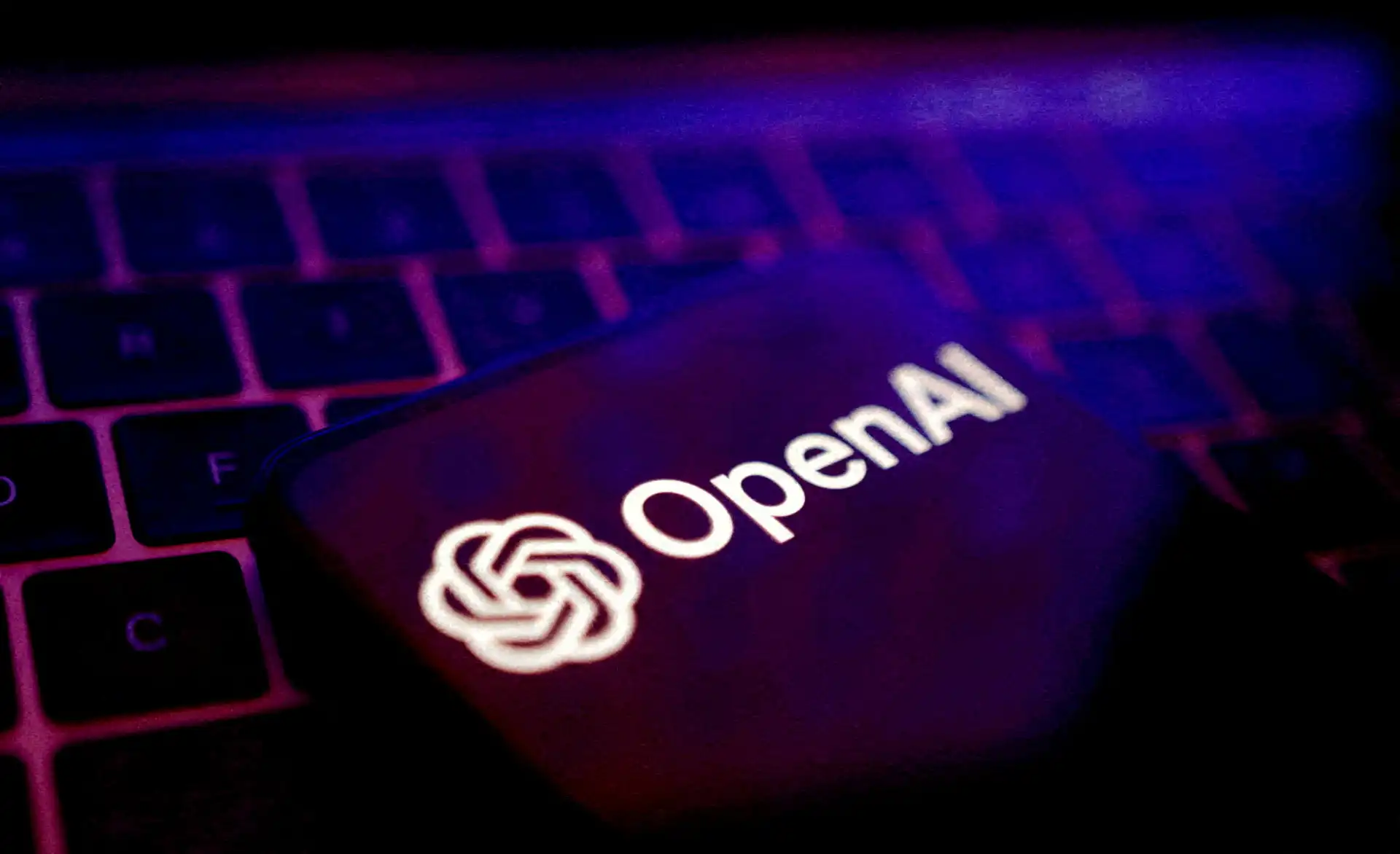 Dispositivo móvel exibindo o logo da OpenAI com um teclado de computador ao fundo, iluminado por luzes roxas e azuis, simbolizando tecnologia e inovação em inteligência artificial.