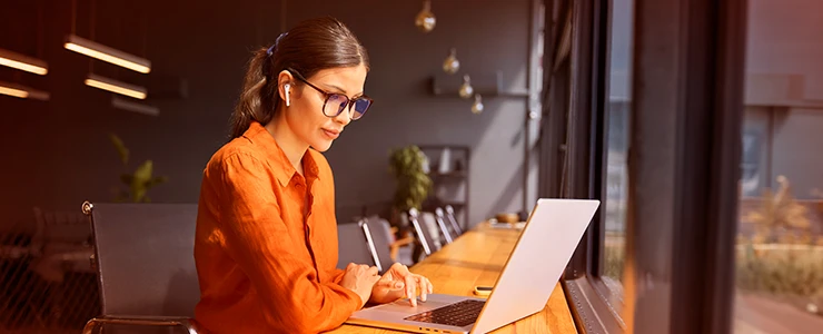 Mulher de óculos e cabelo preso trabalhando em um notebook em um ambiente moderno com iluminação quente e vista para o pôr do sol. direito publicitário