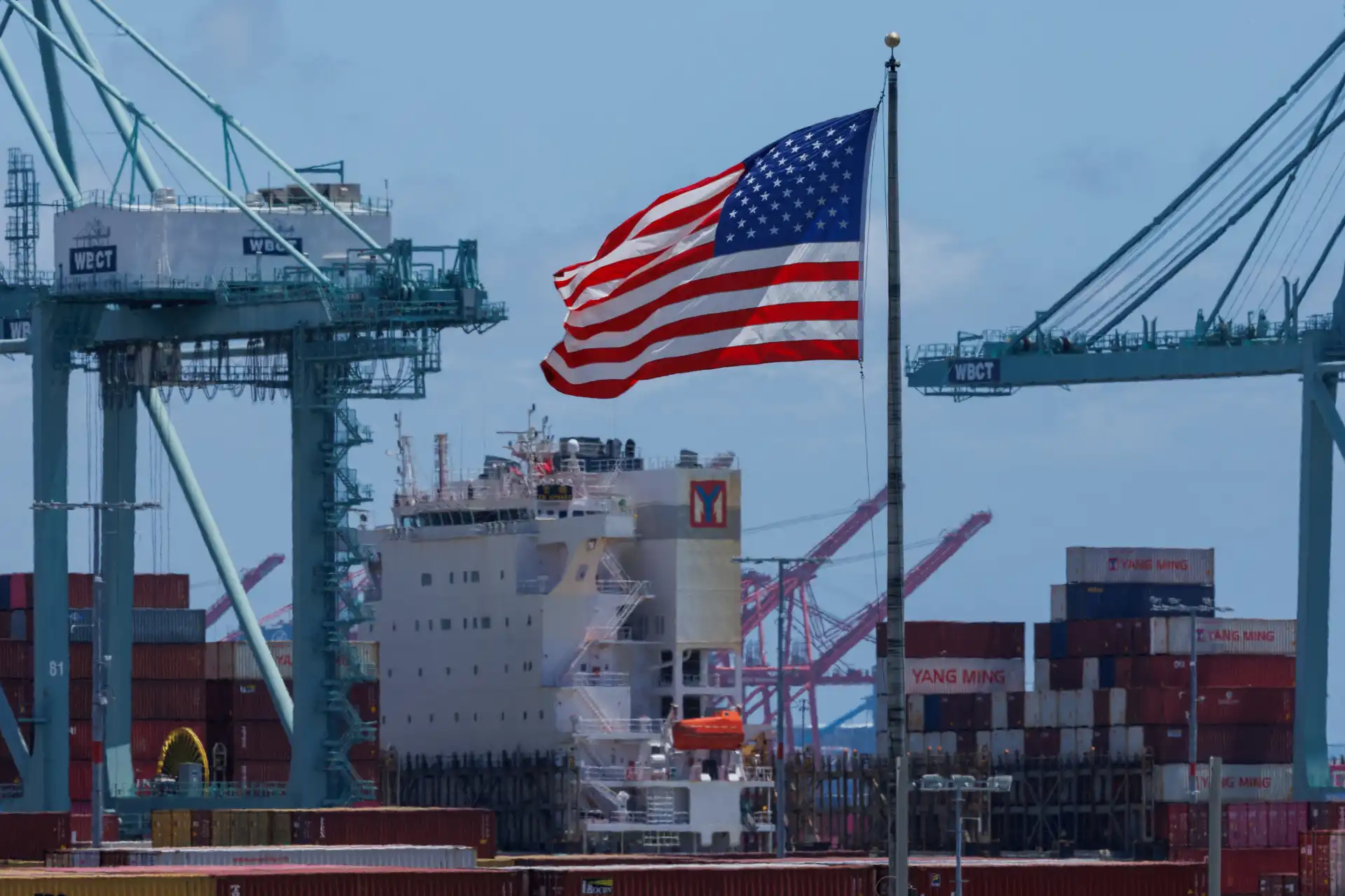 Imagem de um navio de carga no porto com bandeira dos Estados Unidos ao vento e containers ao fundo, simbolizando comércio internacional e transporte marítimo.
