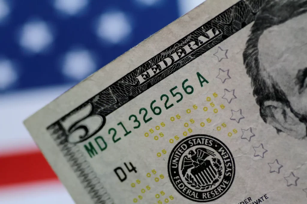 Imagem de uma nota de cinco dólares dos Estados Unidos com foco na parte superior, destacando o número 5 e detalhes de segurança, com fundo desfocado.