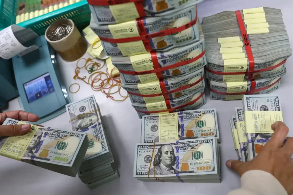 Imagem de várias pilhas de dinheiro em dólares americanos sendo contadas na mesa, com fita vermelha e amarela ao redor das pilhas, garantindo segurança e organização.