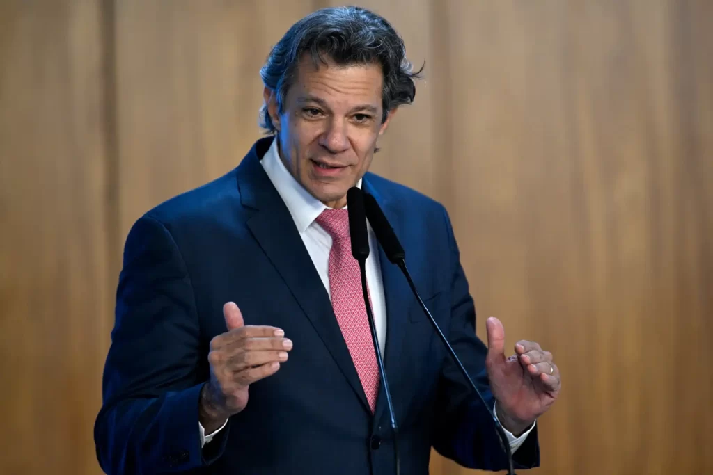 Imagem de um político brasileiro falando em um evento, vestido com terno azul, camisa branca e gravata rosa, com fundo de painel de madeira, representando pauta de política e liderança.
