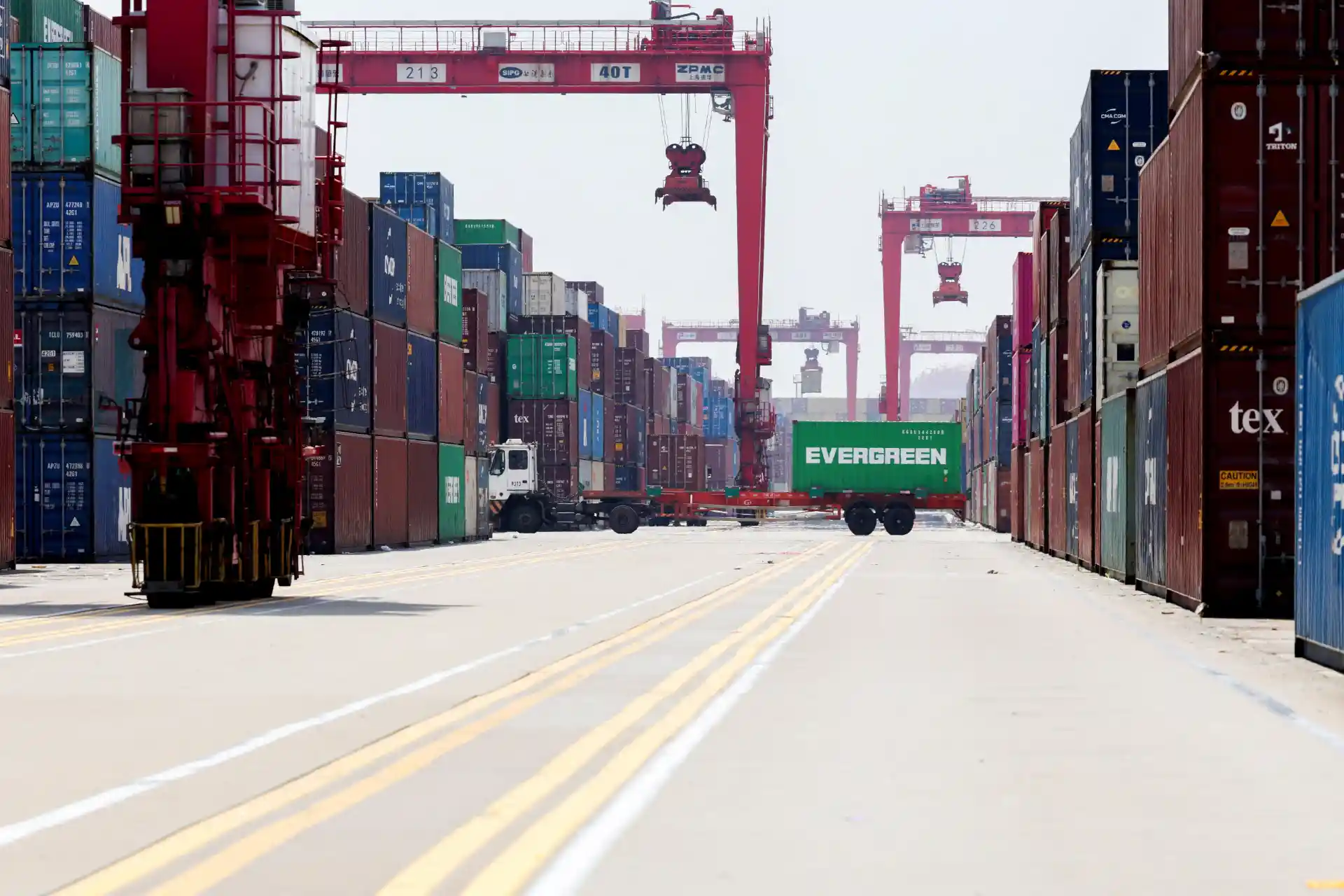 Imagem de um porto de carga com contêineres coloridos empilhados, guindastes de carga e caminhões, representando logística e transporte internacional.