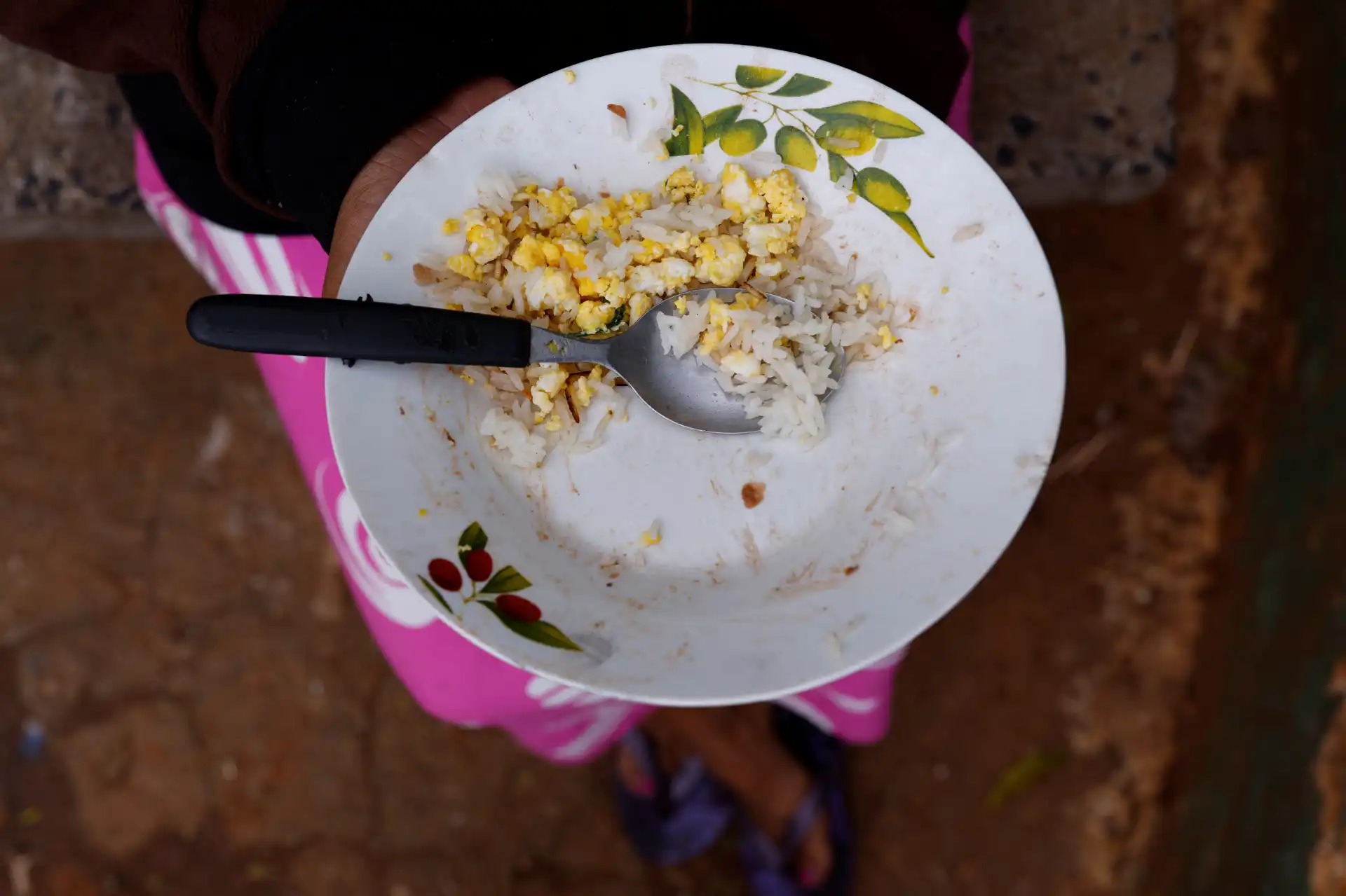 Prato com arroz, ovo mexido e uma colher, sustentado por uma criança com calça rosa e sapatos pretos, ao ar livre sobre solo de terra.