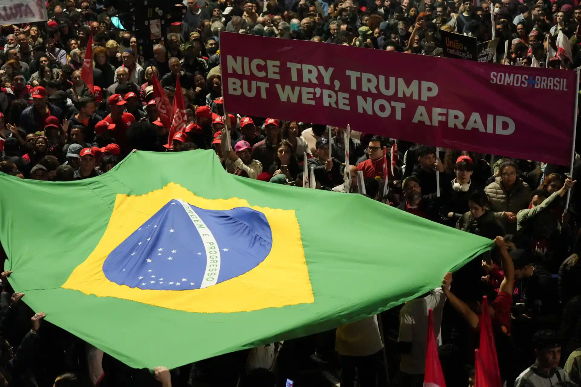 Protesto no Brasil com bandeira brasileira grande e pessoas segurando cartaz que diz "Nice try, Trump but we're not afraid" em uma manifestação de apoio ao país.
