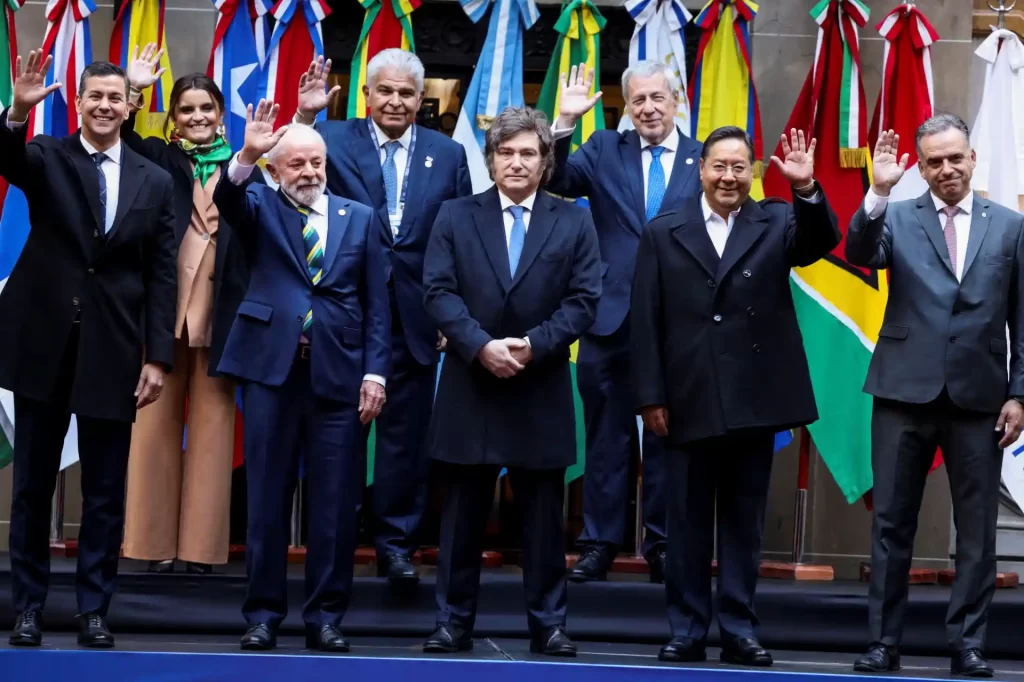Reunião de líderes internacionais com bandeiras de países ao fundo, todos vestindo trajes formais e acenando para a câmera, promovendo diplomacia e cooperação mundial.