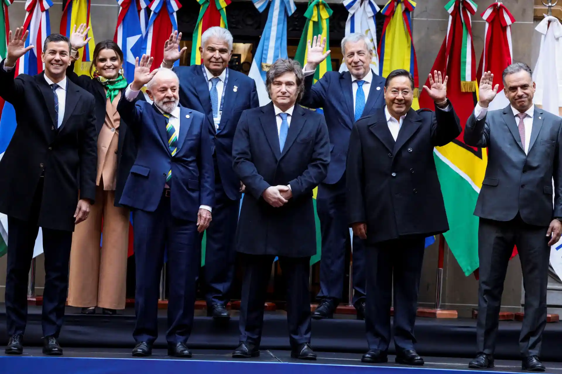 Reunião de líderes internacionais com bandeiras de países ao fundo, todos vestindo trajes formais e acenando para a câmera, promovendo diplomacia e cooperação mundial.