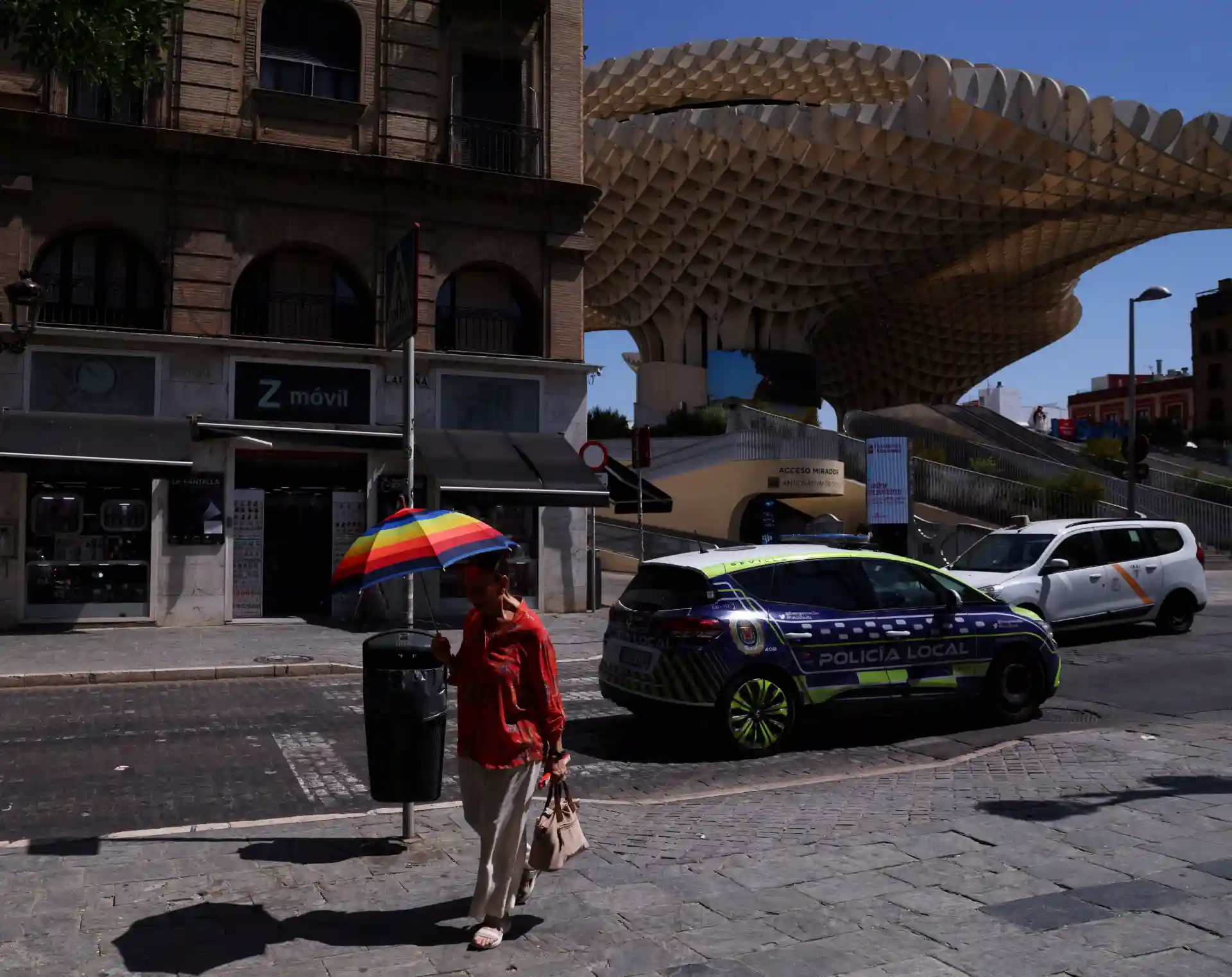 Imagem de uma rua em Granada, Espanha, com um policial local, uma pessoa com guarda-chuva colorido, carros em movimento e a arquitetura moderna e histórica ao fundo.