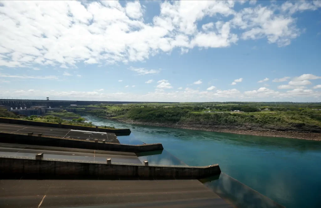 Imagem da usina hidrelétrica com reservatório de água e landscape ao fundo, sob céu parcialmente nublado, simbolizando energia renovável.