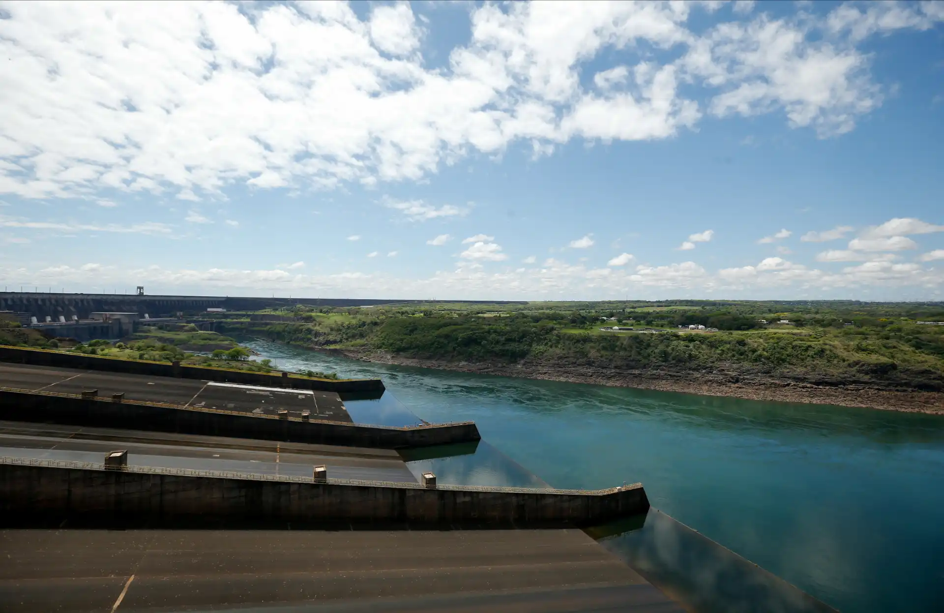 Imagem da usina hidrelétrica com reservatório de água e landscape ao fundo, sob céu parcialmente nublado, simbolizando energia renovável.