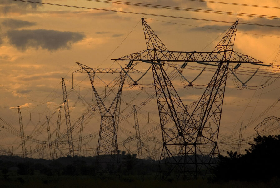 Imagem de torres de transmissão de energia elétrica ao pôr do sol, com céu alaranjado ao fundo, destacando a infraestrutura de distribuição de energia.