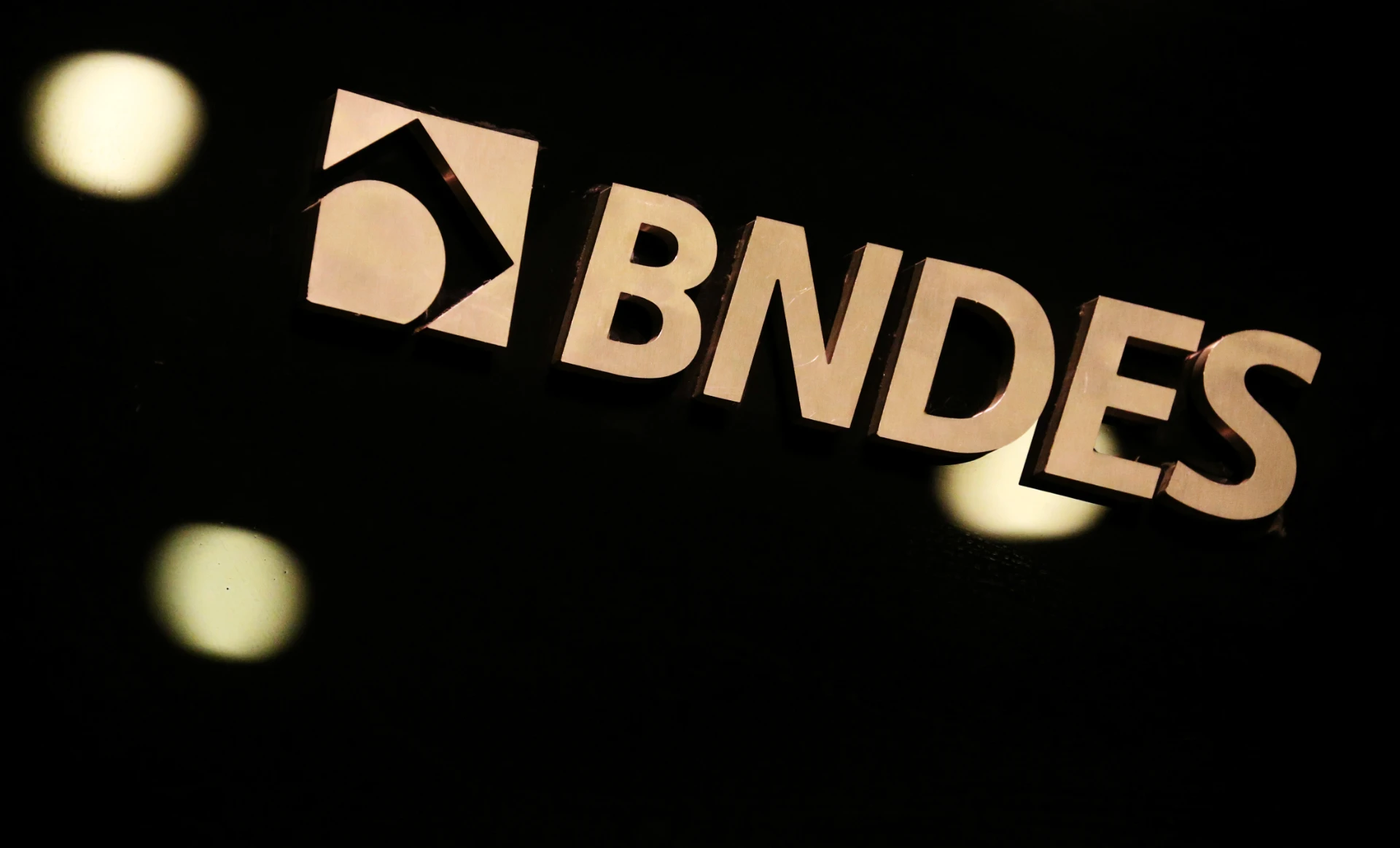 Imagem da logomarca do BNDES iluminada em um ambiente escuro, destacando o nome e o símbolo do banco de desenvolvimento brasileiro.