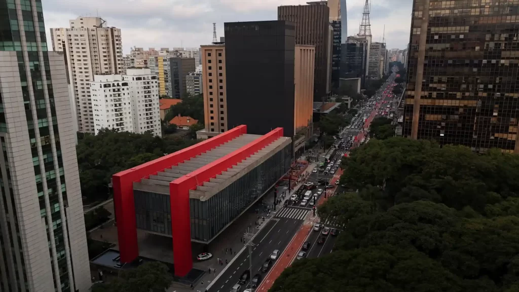 Vista de um centro urbano moderno com prédios altos e movimentado fluxo de veículos na rua, destacando o edifício com estrutura vermelha.