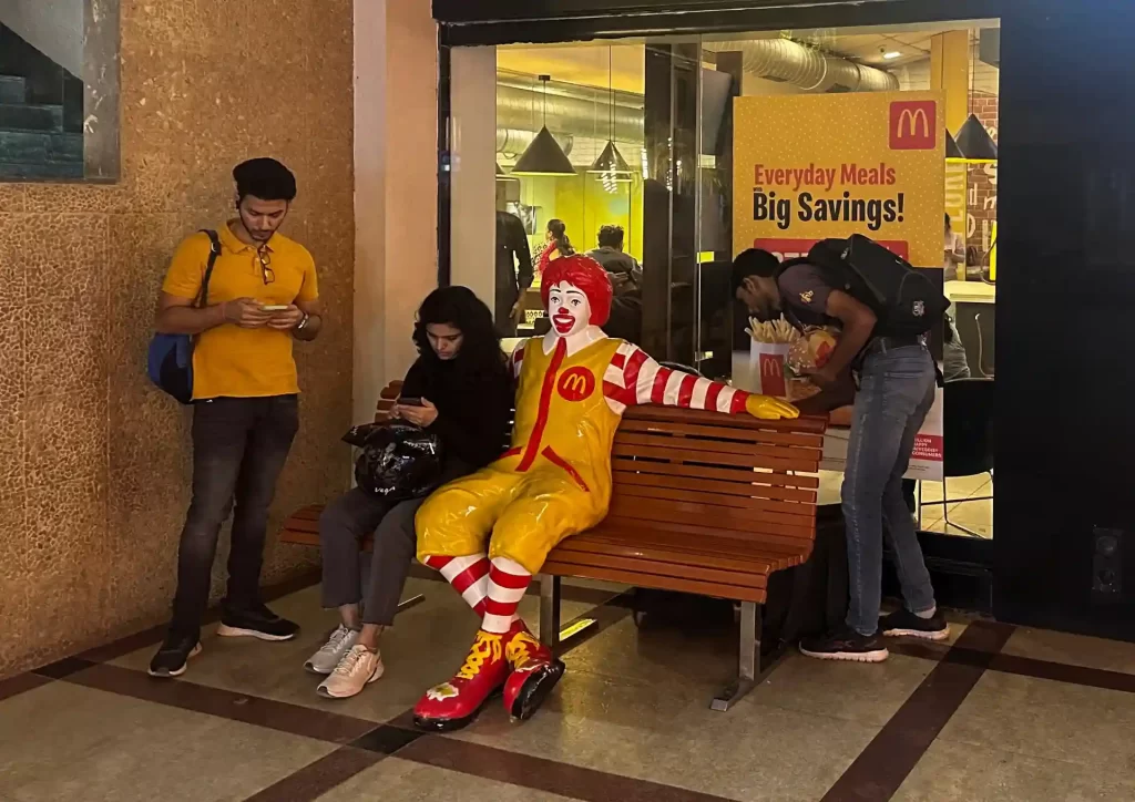 Clientes esperando no McDonald's com um Ronald McDonald vivo na praça de alimentação, na frente de uma fachada com promoção de refeições diárias e economia.
